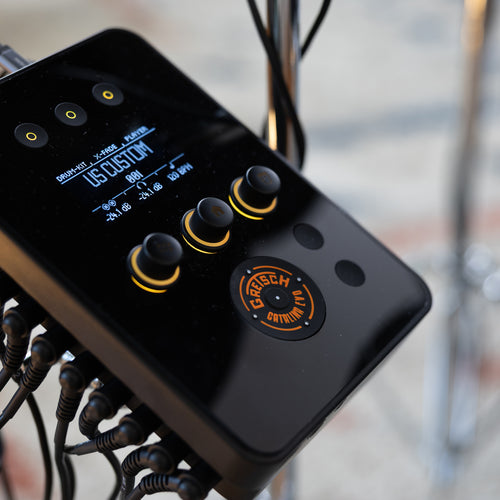 A close-up of a black electronic drum module with multiple knobs, a small blue display screen, and attached cables, photographed in a real studio or performance environment with blurred drum hardware in the background.