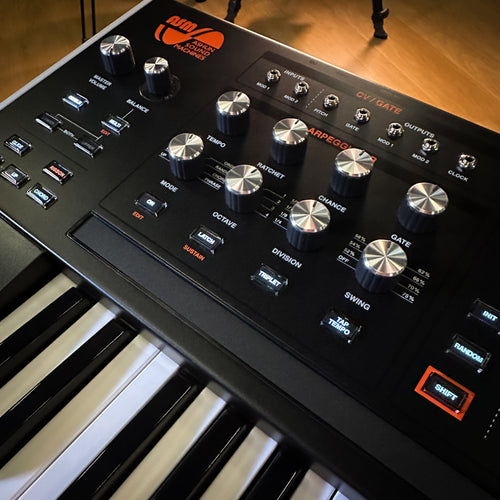 A close-up, angled view of a ASM Hydrasynth Deluxe Polyphonic Wavemorphing Synthesizer, showing its black faceplate with silver and black knobs, white and black piano keys, and orange branding text against a warm wooden floor background.