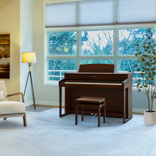 Kawai CA501 Concert Artist Digital Piano - Mocha Walnut in a warm brown wood finish, shown in a bright, modern living room setting with a matching bench, a floor lamp, and large windows looking out to trees.