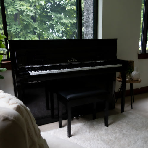 A Kawai NOVUS NV6 Hybrid Upright Piano in a home interior setting with large windows showing green foliage outside.
