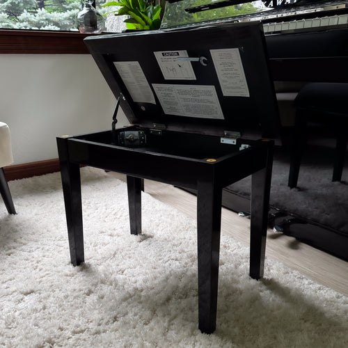 A Kawai piano bench, open with a padded black seat cushion, positioned on a white shag rug next to a Kawai NOVUS NV6 Hybrid Upright Piano.