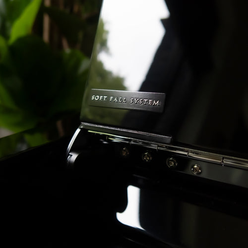 Close-up of a Kawai NOVUS NV6 Hybrid Upright Piano lid with the words 'SOFT FALL SYSTEM' embossed on a metal plate, showing hinges and screws against a dark background with blurred green foliage.