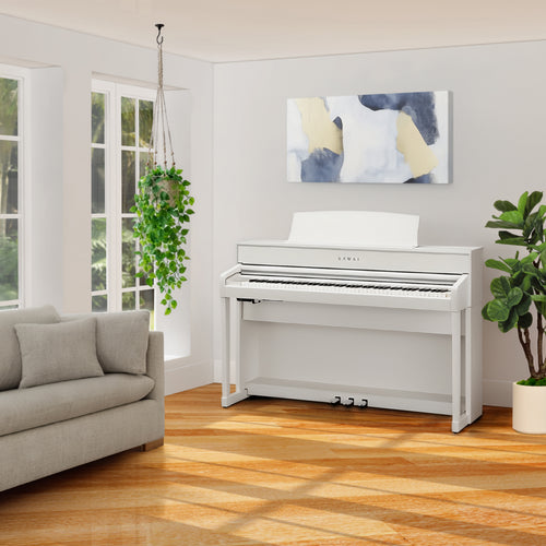 Kawai CA701 Digital Piano in Satin White in a bright, modern living room setting with a gray sofa, wooden floors, and a potted plant.