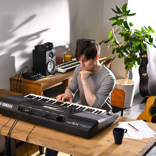 Person playing a Yamaha keyboard in a home studio setting with a laptop, speakers, and musical instruments.