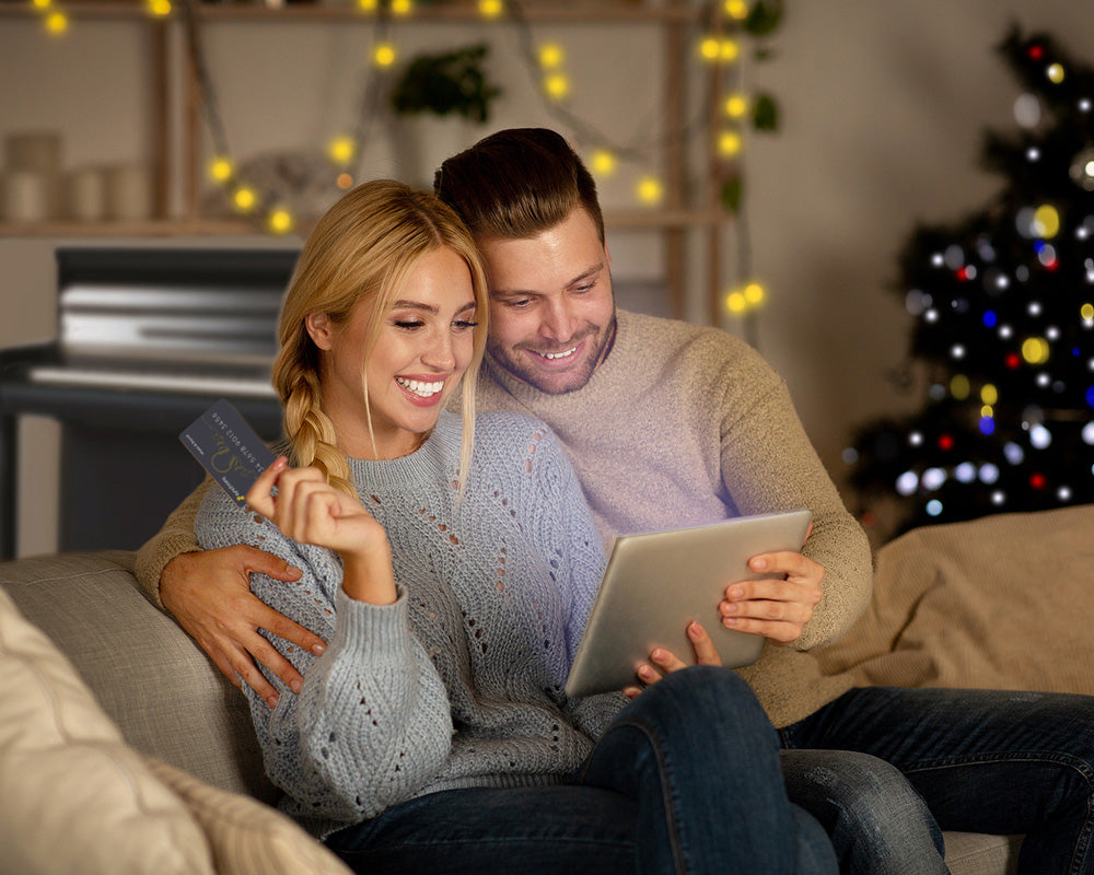 Image of man and woman shopping on iPad with Synchrony credit card, piano and Christmas tree in the background.