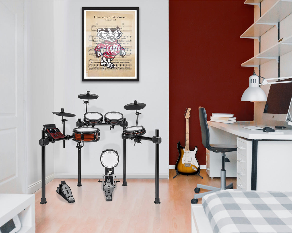 A University of Wisconsin Badgers framed poster hangs on a white wall above an electronic drum kit in a modern room with a red accent wall, desk, guitar, and computer setup.