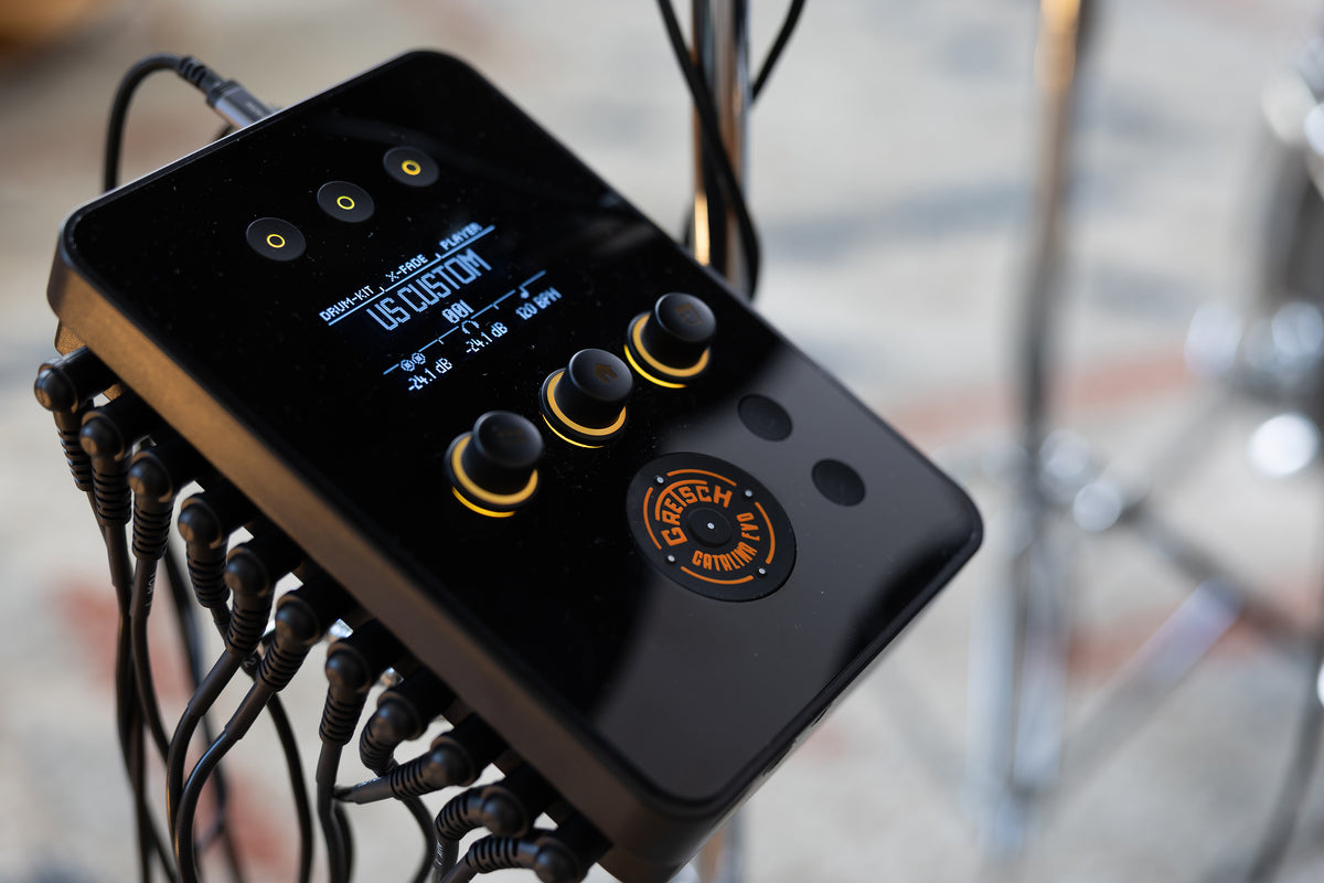 A close-up of a black electronic drum module with multiple knobs, a small blue display screen, and attached cables, photographed in a real studio or performance environment with blurred drum hardware in the background.