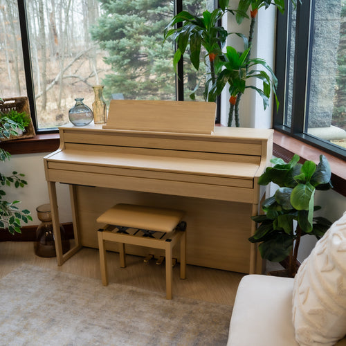 Roland HP704 Digital Piano - Light Oak with a bench in a room with large windows and plants, with the key cover closed