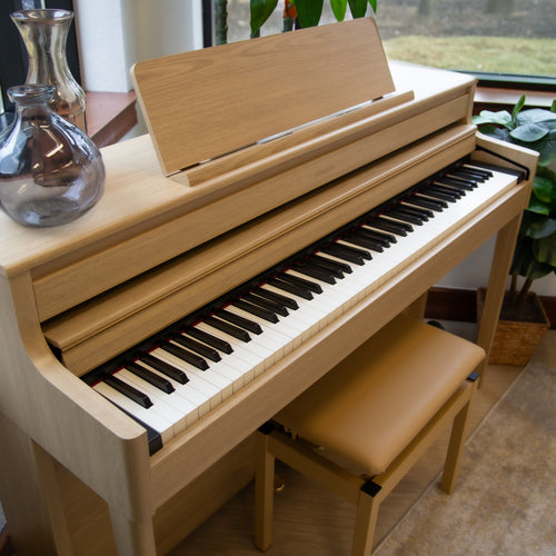 Roland HP704 Digital Piano - Light Oak with a bench in a room with plants and a window, showing the key cover partially closed to hide controls