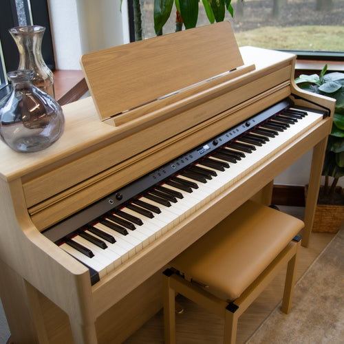 Roland HP704 Digital Piano - Light Oak with a bench in a room with plants and a window, showing the key cover open to show controls