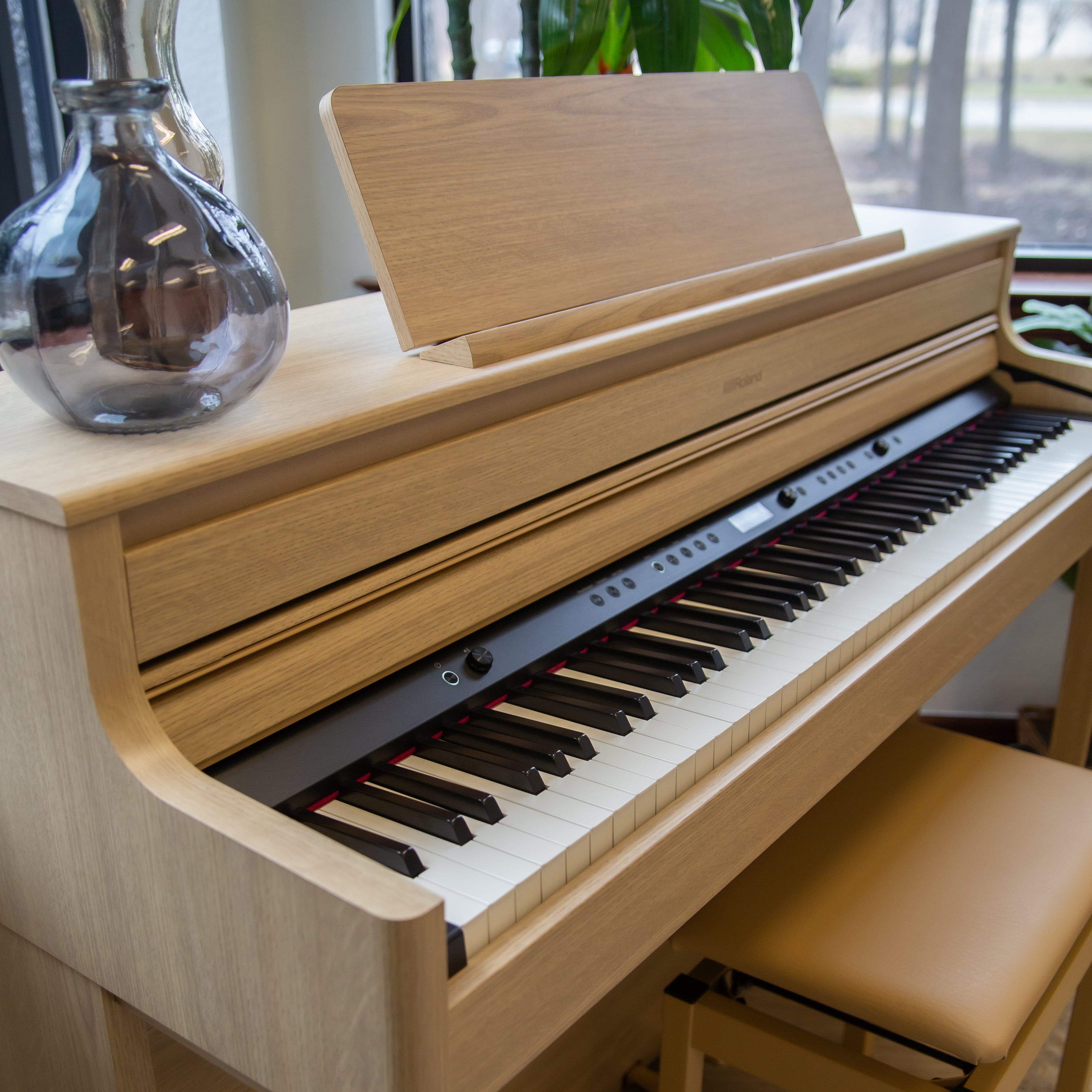 Roland HP704 Digital Piano - Light Oak with a clear glass vase on top, set against a blurred indoor background.