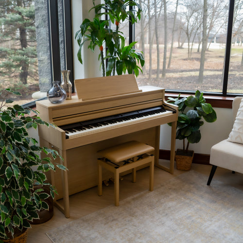 Roland HP704 Digital Piano - Light Oak in a room with large windows and indoor plants