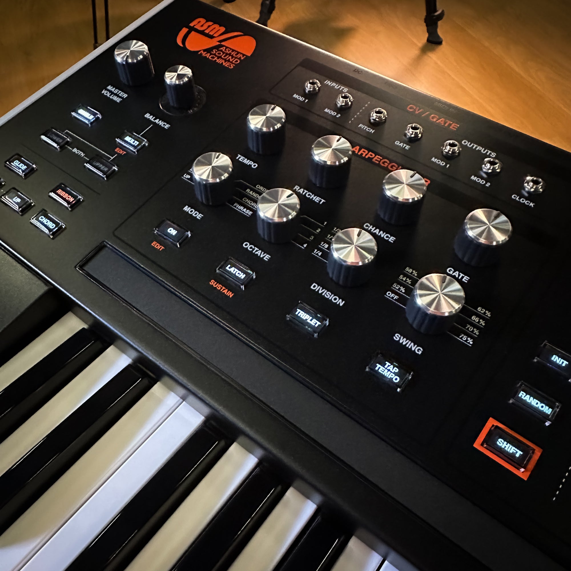A close-up, angled view of a ASM Hydrasynth Deluxe Polyphonic Wavemorphing Synthesizer, showing its black faceplate with silver and black knobs, white and black piano keys, and orange branding text against a warm wooden floor background.