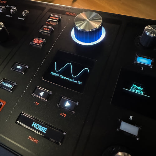 Close-up of a ASM Hydrasynth Deluxe Polyphonic Wavemorphing Synthesizer with illuminated buttons, a large silver-and-blue rotary knob, and a small display showing a waveform.