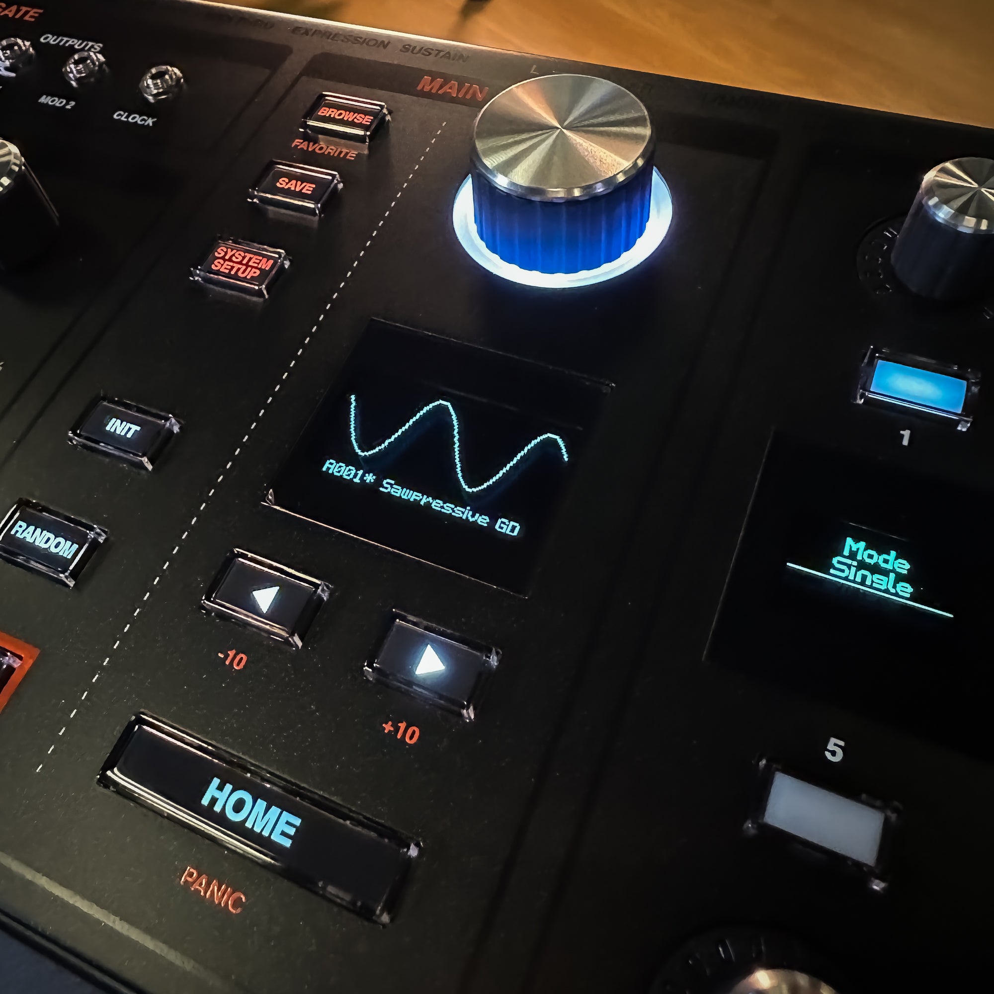 Close-up of a ASM Hydrasynth Deluxe Polyphonic Wavemorphing Synthesizer with illuminated buttons, a large silver-and-blue rotary knob, and a small display showing a waveform.