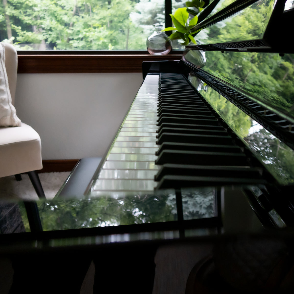 A close-up, angled view of a Kawai NOVUS NV6 Hybrid Upright Piano, set in a room with a window showing lush green trees outside.