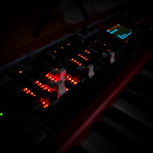 A close-up, angled view of a Nord Electro 7 with illuminated display screen, set against a very dark background.