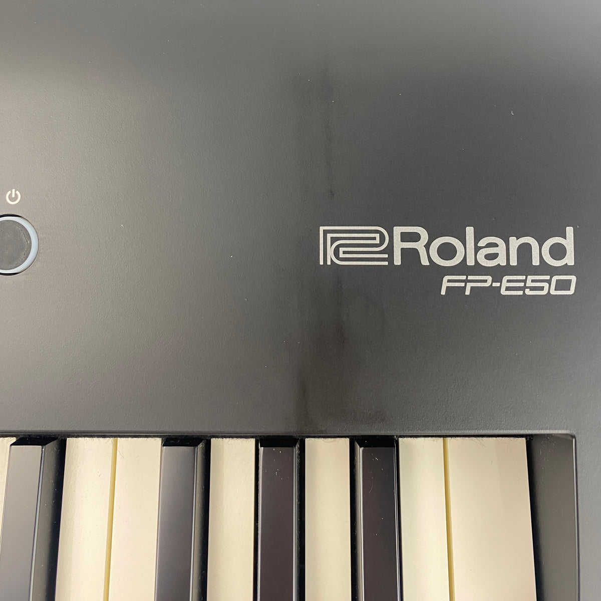 Close-up of Roland FP-E50 logo and power button on matte black panel with light smudges and scuffing