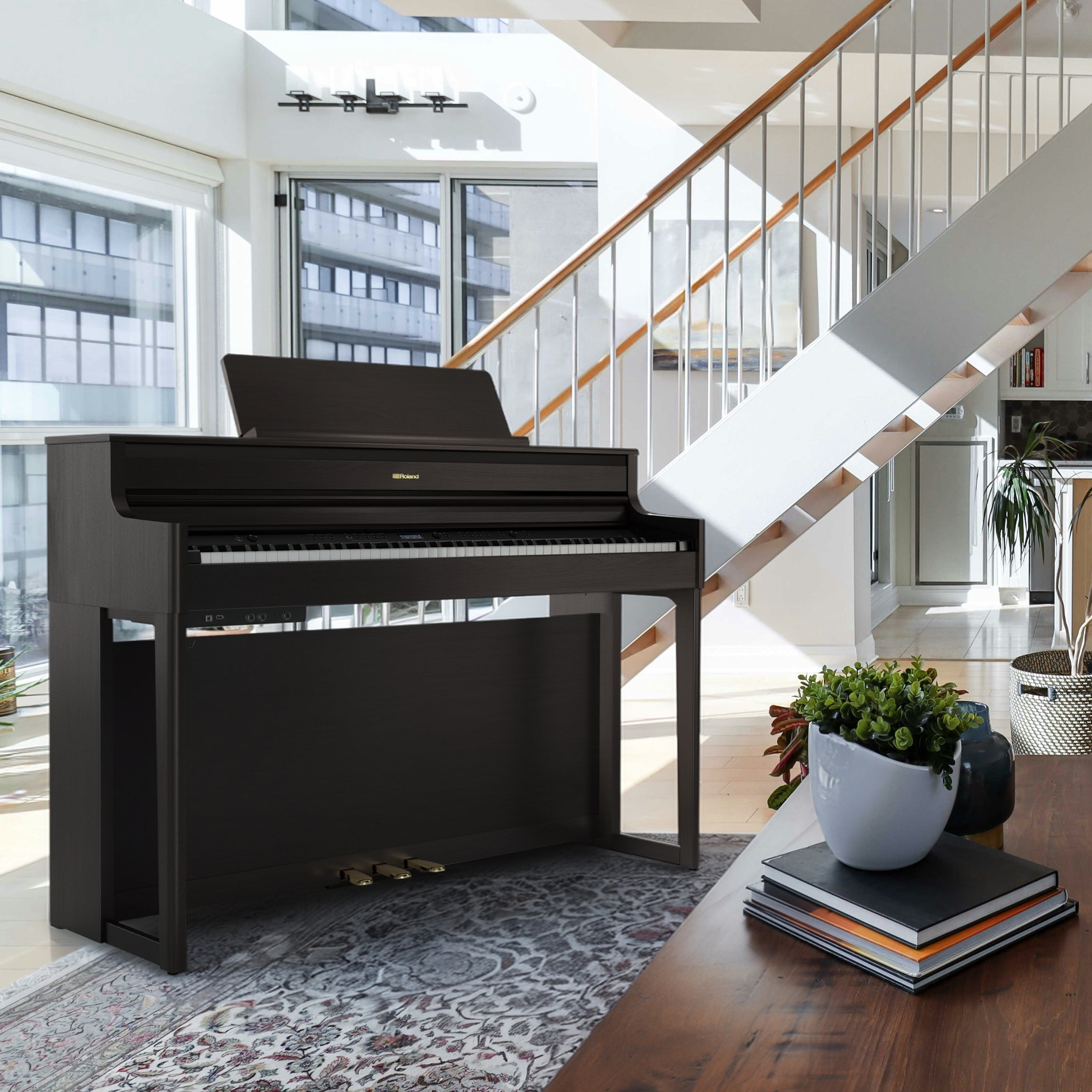 Roland HP704 Digital Piano - Dark Rosewood - in a modern living room with a staircase and large windows.