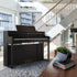 Roland HP704 Digital Piano - Dark Rosewood - in a modern living room with a staircase and large windows.