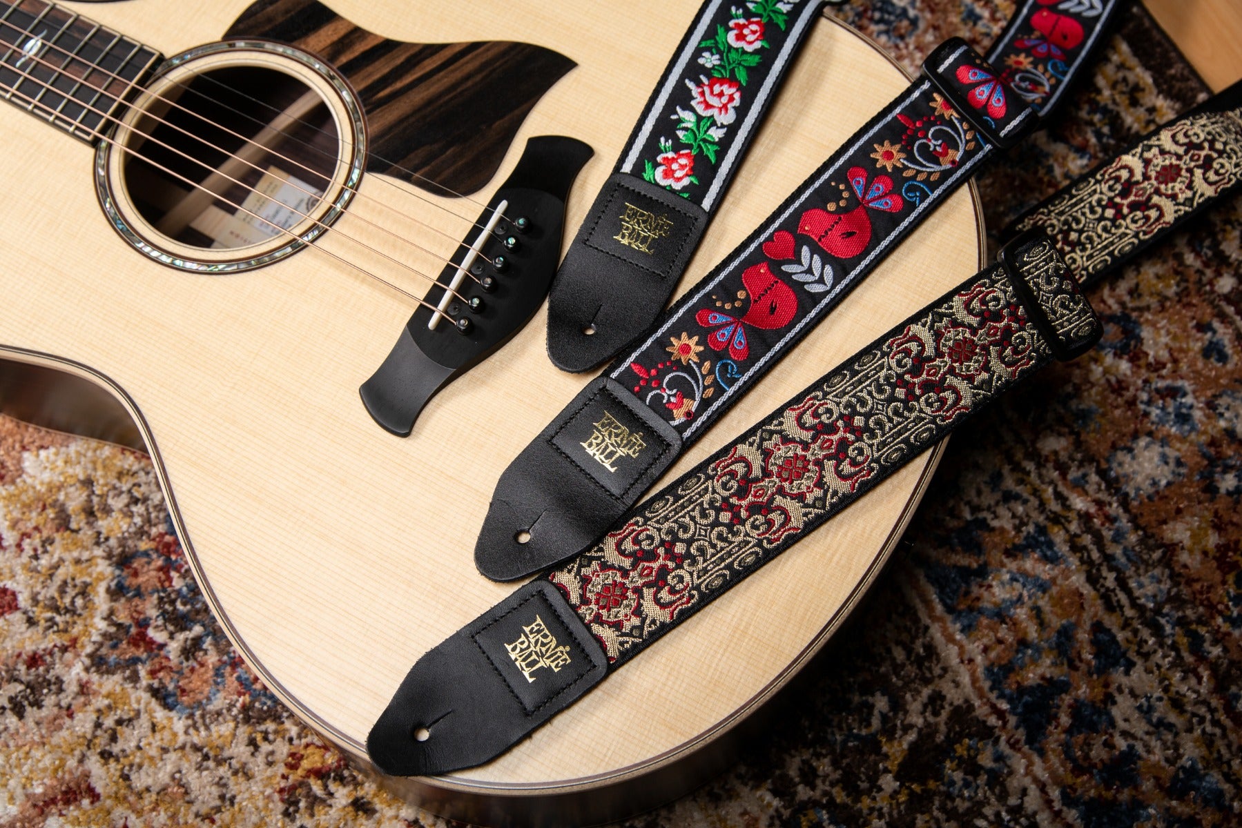 Image of several guitar straps including the Ernie Ball Persian Gold Jacquard Guitar Strap laying on the body of a Taylor guitar